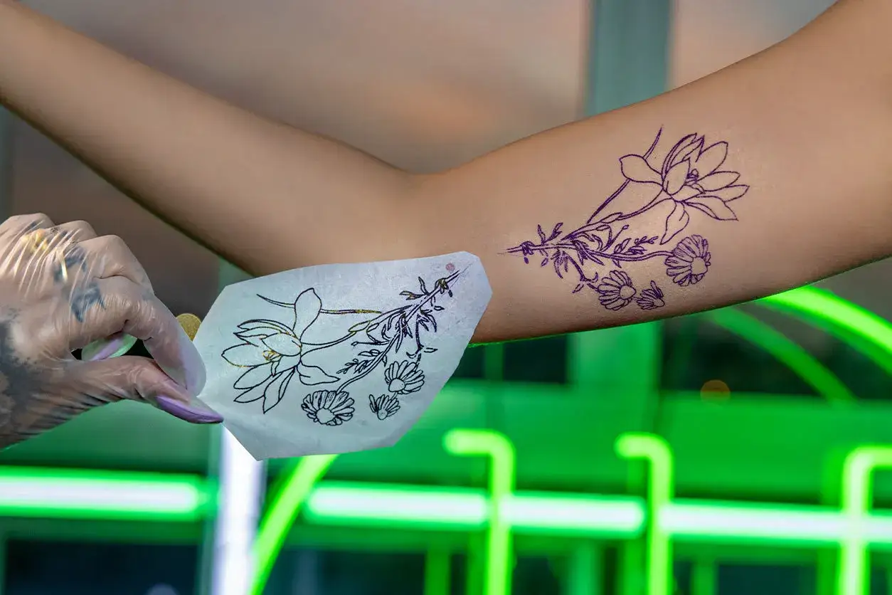 Manos enguantadas sostienen una plantilla de tatuaje floral. El diseño se alinea con un tatuaje temporal ya en el brazo, listo para la aplicación de crema para antes de tatuarse.