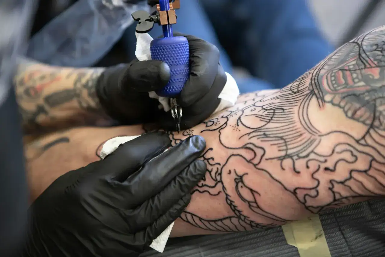 Manos enguantadas trabajan con m&aacute;quina de tatuar, aplicando la mejor tinta para tatuajes en un brazo con dise&ntilde;os de drag&oacute;n y olas.