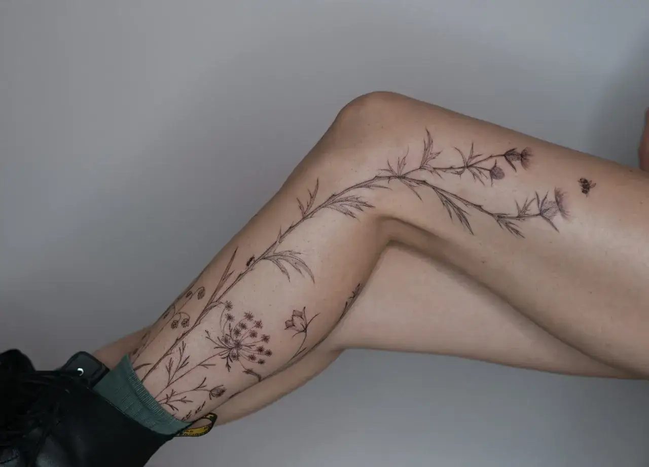 Tatuaje de ramas y flores silvestres en la pierna de Sara Calixto, con una abeja y una hormiga.