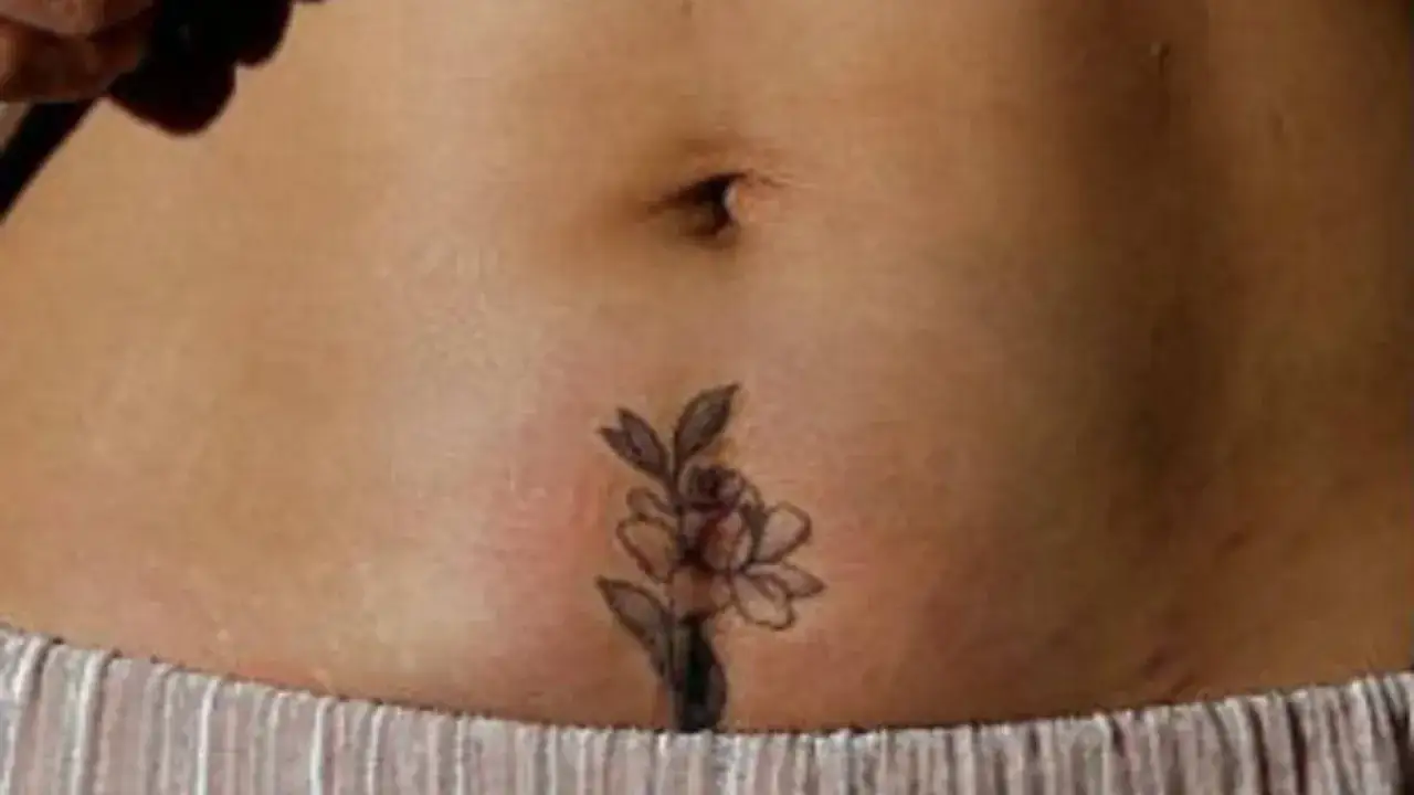 Un delicado tatuaje de flor adorna la piel de una mujer, justo debajo de su ombligo, cubriendo sutilmente una cicatriz.