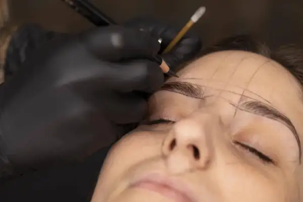 Manos enguantadas dibujan cejas con lápiz, marcando el rostro de Jessica Wilde para un diseño preciso.