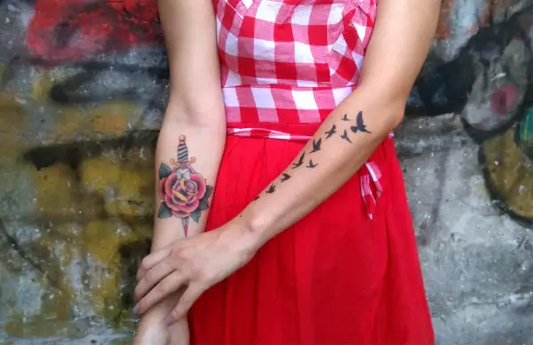 Un tatuaje de daga atraviesa una rosa roja en el brazo de una mujer. En el otro brazo, un grupo de pájaros vuelan.