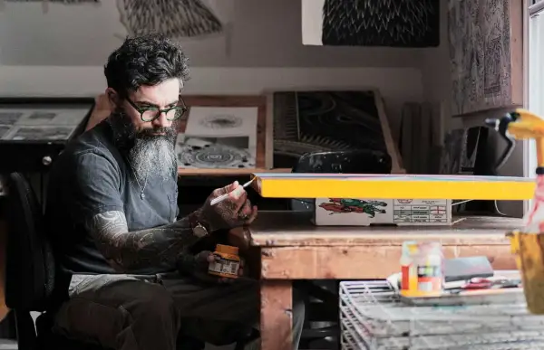 Thomas Hooper, con barba y gafas verdes, pinta un objeto amarillo en su estudio.
