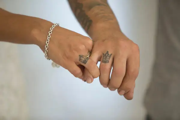 Tatuaje manos entrelazadas significado: dos coronas en los dedos, símbolo de unión y realeza compartida.