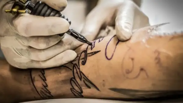 Manos enguantadas crean un diseño de letra con una máquina de tatuar. Un tatuaje con buena tinta se está formando en la piel.