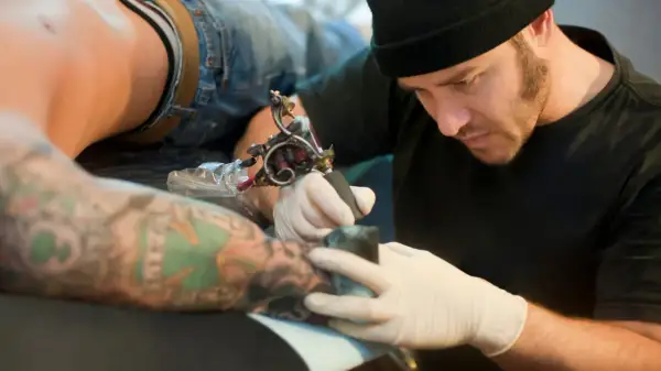 Artista tatuador concentrado, usando la mejor tinta para tatuajes en un brazo con diseños coloridos.