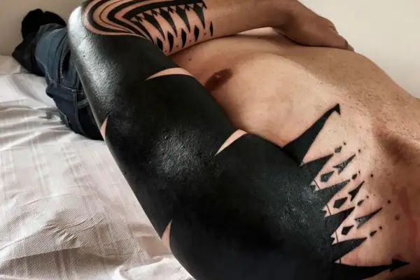 Un hombre con tatuajes blackout cubriendo su brazo y pecho, con diseños geométricos y tribales en negro intenso.
