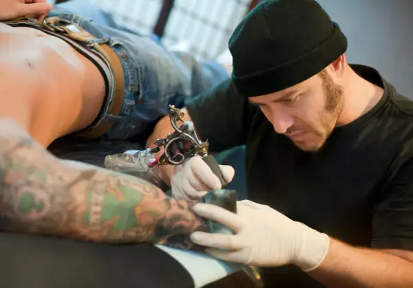 Tatuador con gorro negro y guantes blancos tatuando el brazo de un cliente con tatuajes coloridos.
