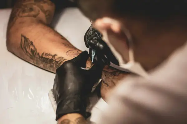 Artista con guantes negros usa máquina para crear un tatuaje en un brazo con diseños previos. La emla para tatuajes ayuda a que el proceso sea más cómodo.