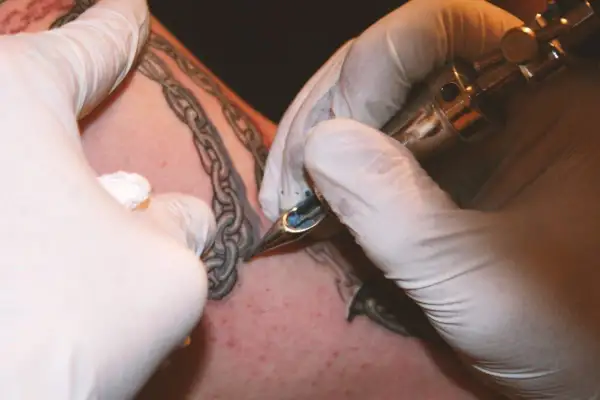 Manos enguantadas tatuando una cadena rota en la piel.
