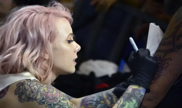 Artista de tatuajes con cabello rosa, con un diseño de megan massacre en su brazo, mientras otro artista trabaja en su piel.