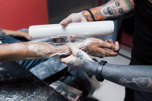 Man wrapping a fresh tattoo with plastic wrap. La curación es clave, y te preguntarás cuántos días hay que ponerse crema en un tatuaje para que quede perfecto.