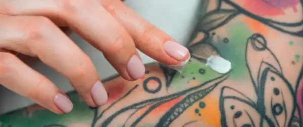 Dedos aplicando crema sobre un tatuaje colorido, parte de un proceso de cuidado y tatuaje terapéutico.