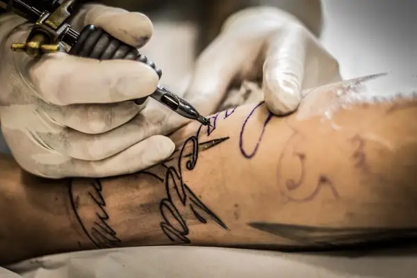 Manos enguantadas trabajan con una máquina de tatuar sobre un brazo, delineando un diseño. Aprende como cuidar un tatuaje recien hecho.