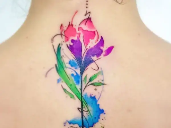 Un vibrante tatuaje de flor estilo acuarela en la espalda, con tonos rosas, morados y verdes. Un diseño de tatuajes de colores para mujer que evoca la naturaleza.