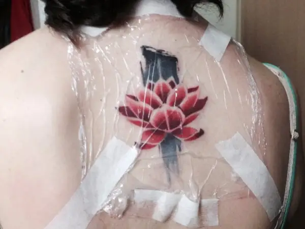 Flor de loto roja y azul recién tatuada en la espalda, cubierta con plástico y cinta. ¿Cuánto se tarda en hacer un tatuaje?
